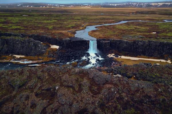 Aerial view of a waterfall cascading into a dark, rocky canyon, surrounded by expansive mossy plains.