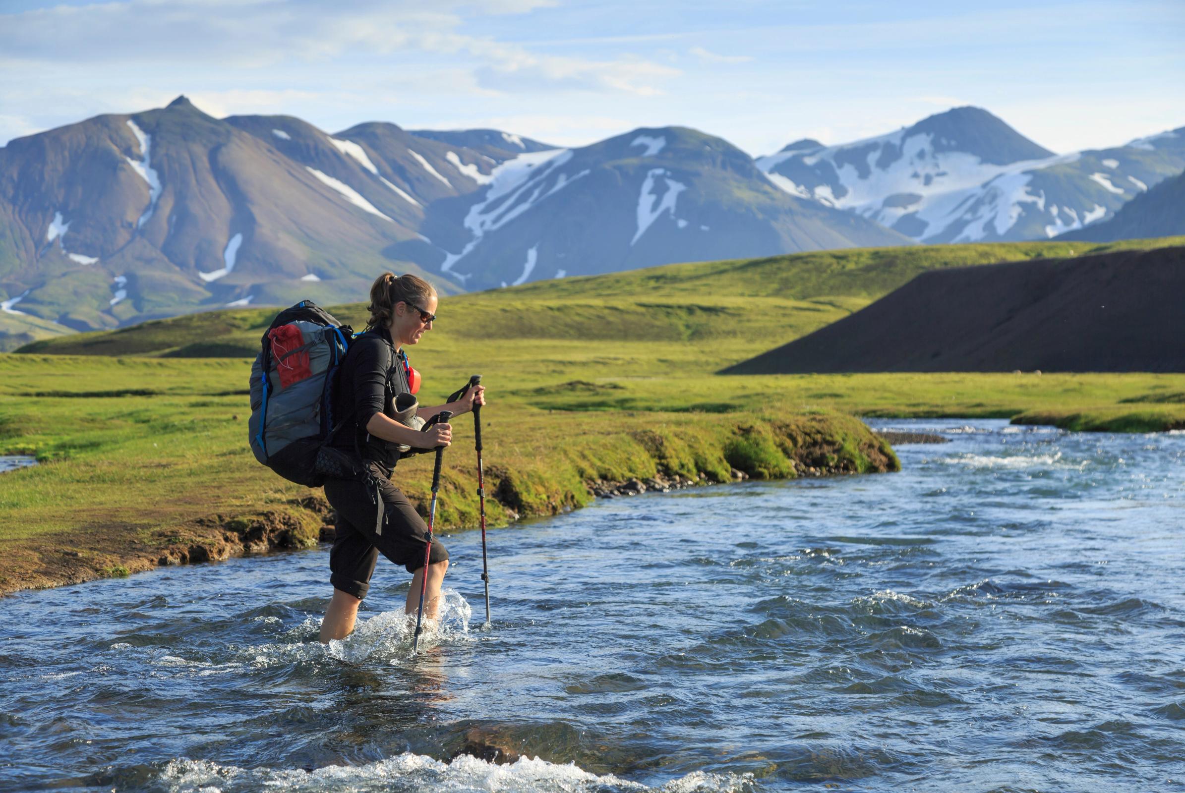 Un excursionista con mochila y bastones de trekking vadea un río en un paisaje montañoso.