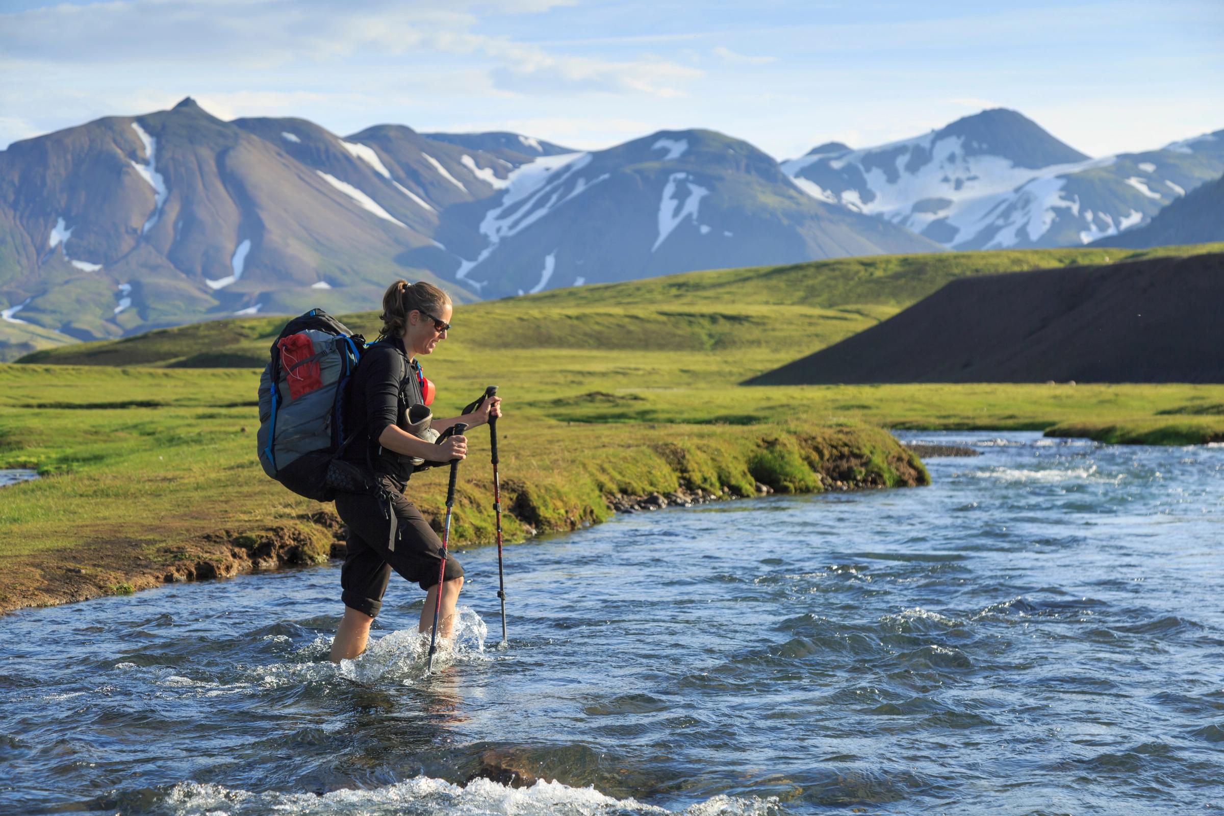 Un excursionista con mochila y bastones de trekking vadea un río en un paisaje montañoso.