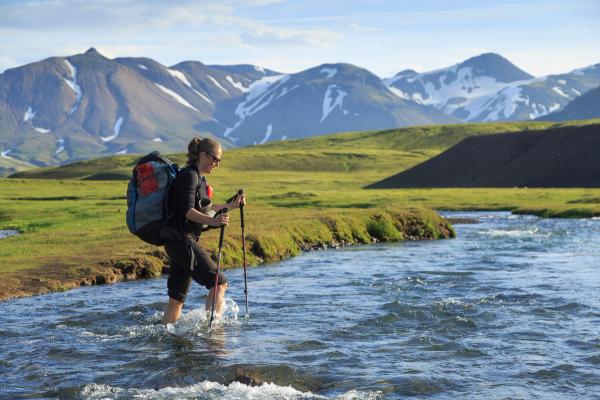 A hiker with a backpack and trekking poles wades through a river in a mountainous landscape.