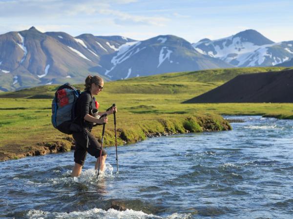 A hiker with a backpack and trekking poles wades through a river in a mountainous landscape.