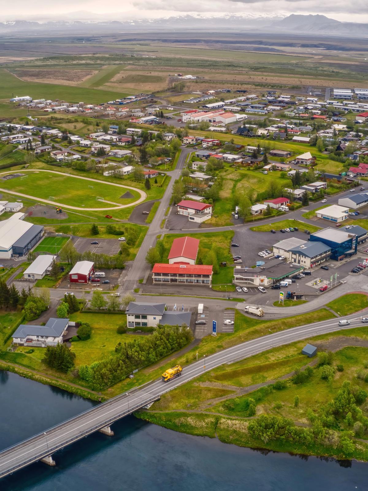 Aerial view of a town with a river and bridge, a sports track, a roundabout, houses, and distant mountains.