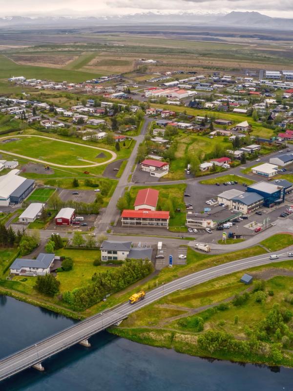 Aerial view of a town with a river and bridge, a sports track, a roundabout, houses, and distant mountains.
