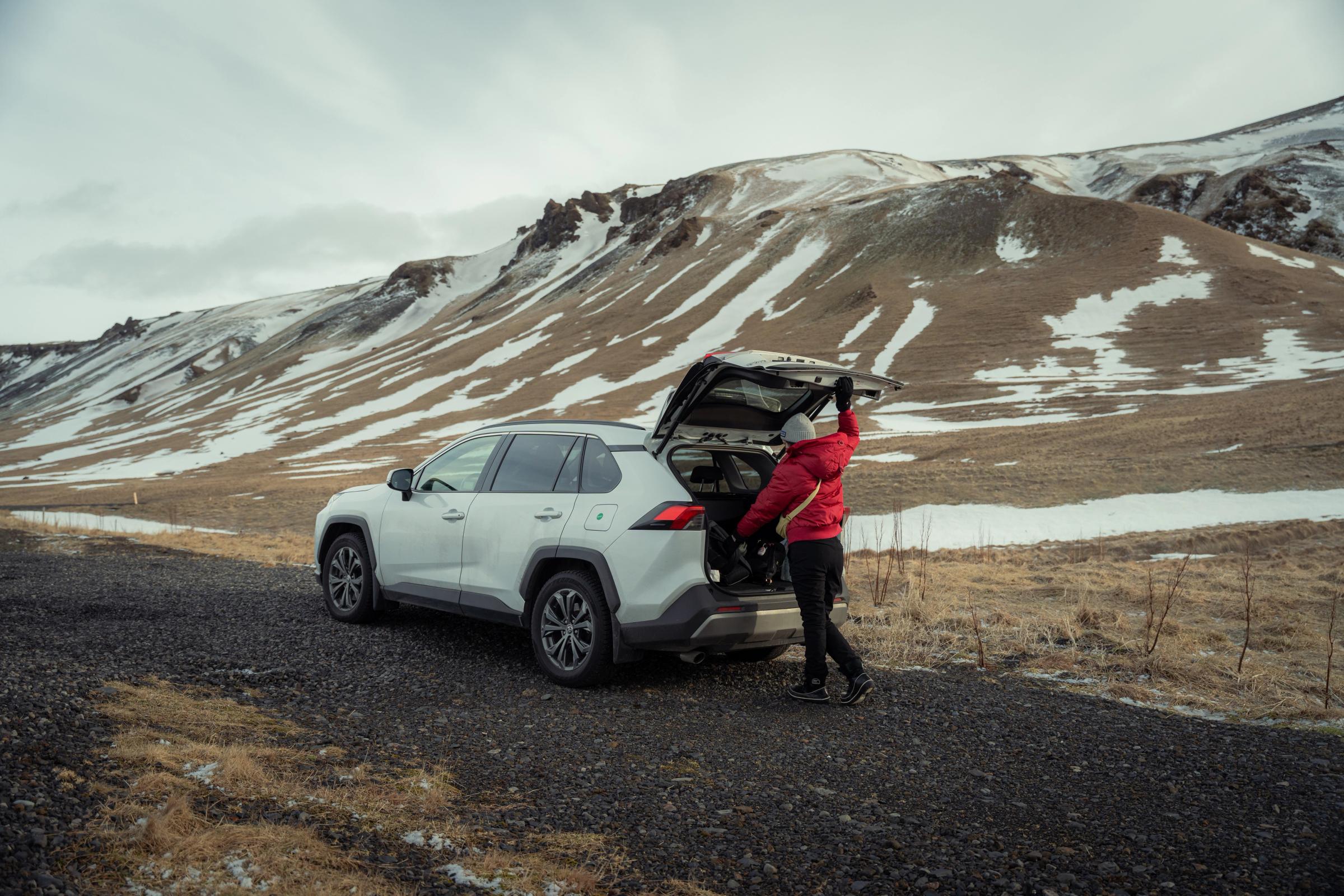 A person loads gear into a white SUV parked on a gravel road amidst snowy mountains.
