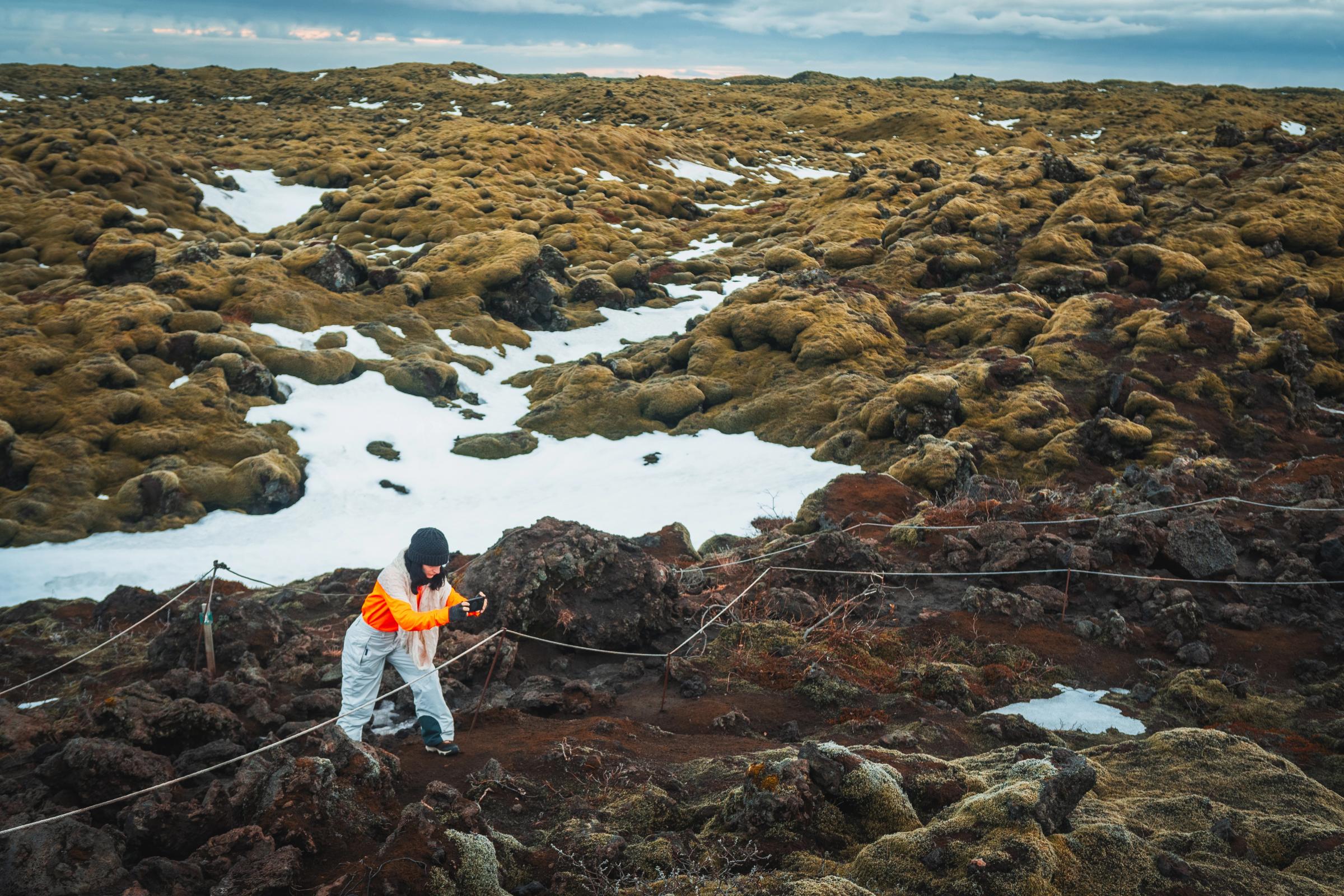 Persona tomando una foto en un extenso campo de lava cubierto de musgo con parches de nieve, detrás de una barrera de cuerda.