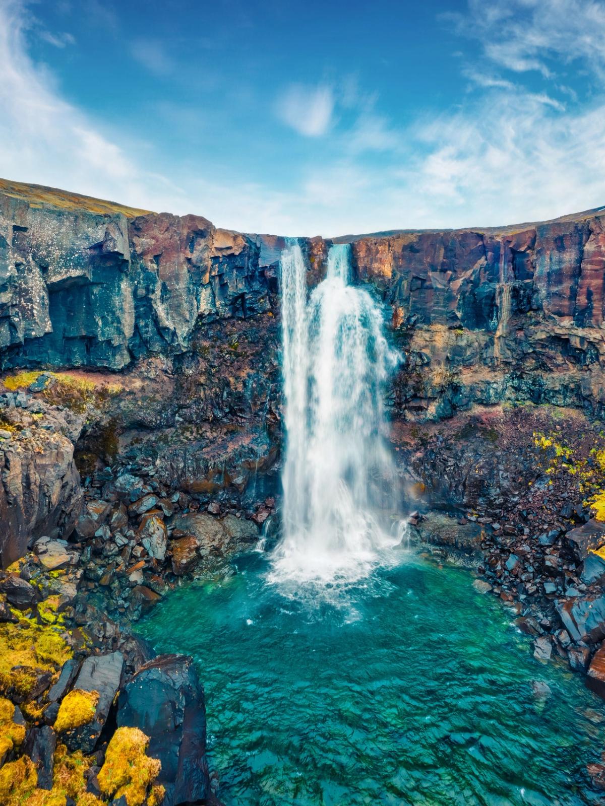 A tall waterfall cascades into a bright turquoise pool surrounded by dark, rugged cliffs, with a person in a red jacket on the right.