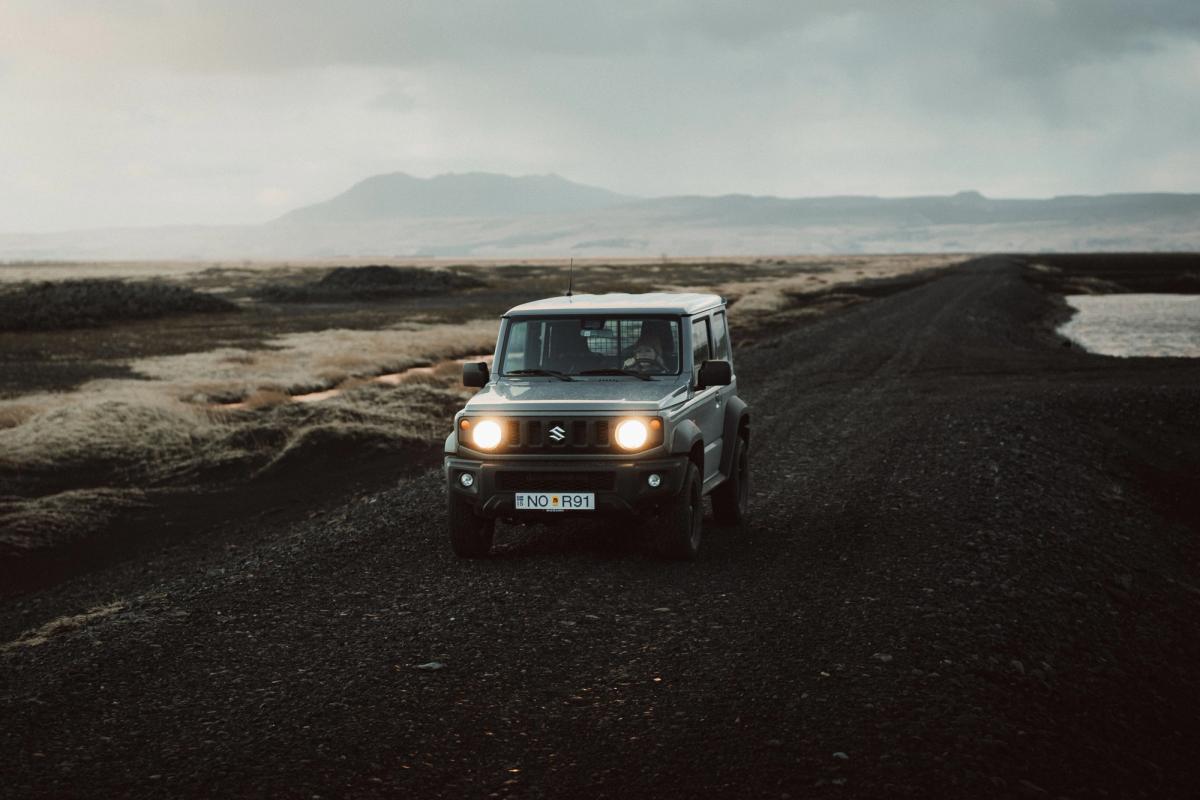 A gray Suzuki Jimny with headlights on drives on a gravel road through a rugged, cloudy landscape.