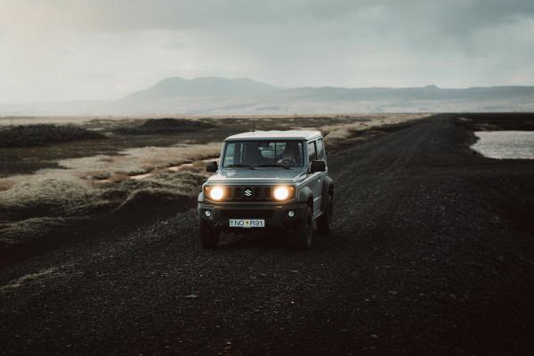 A gray Suzuki Jimny with headlights on drives on a gravel road through a rugged, cloudy landscape.