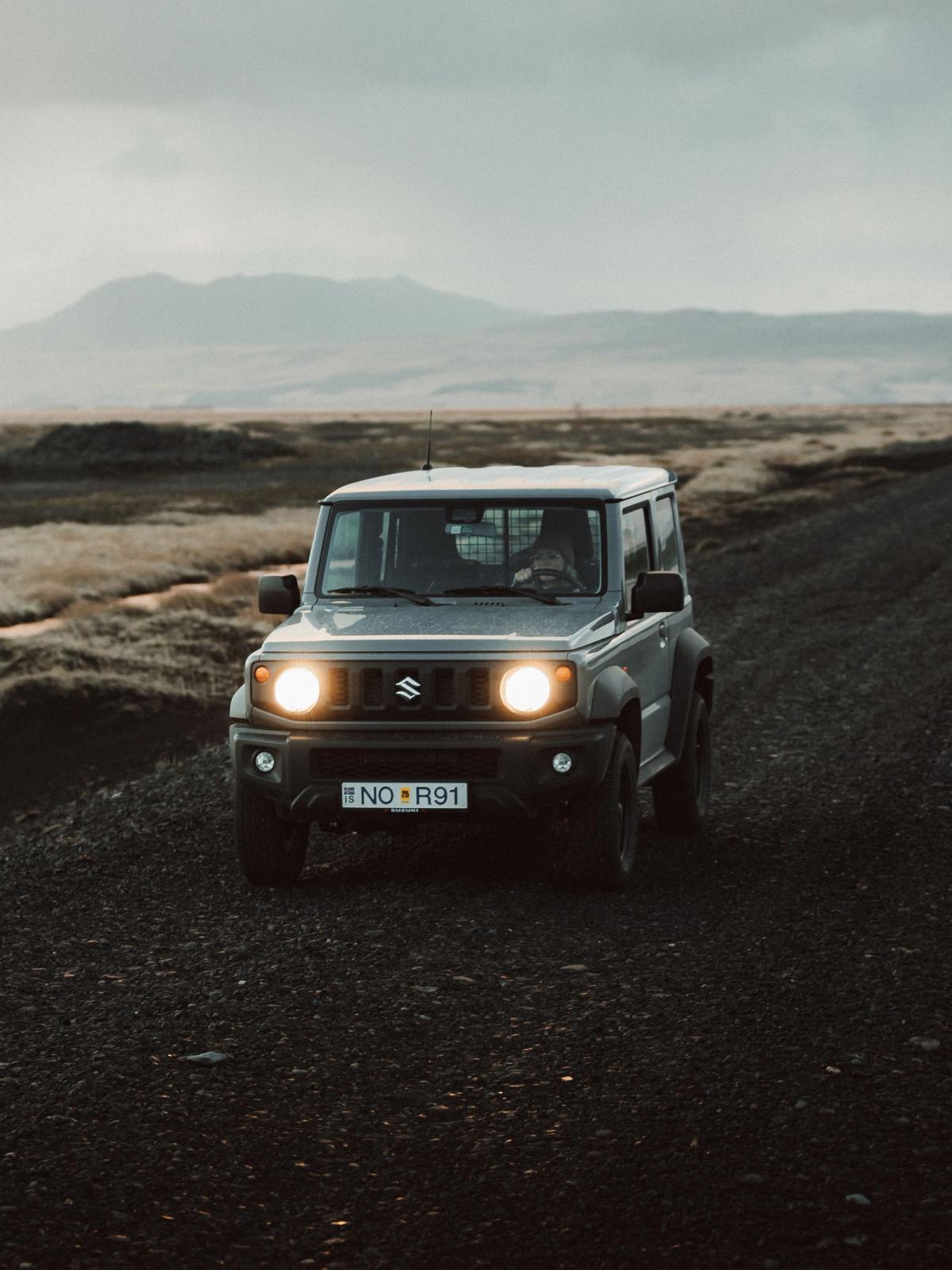 A gray Suzuki Jimny with headlights on drives on a gravel road through a rugged, cloudy landscape.