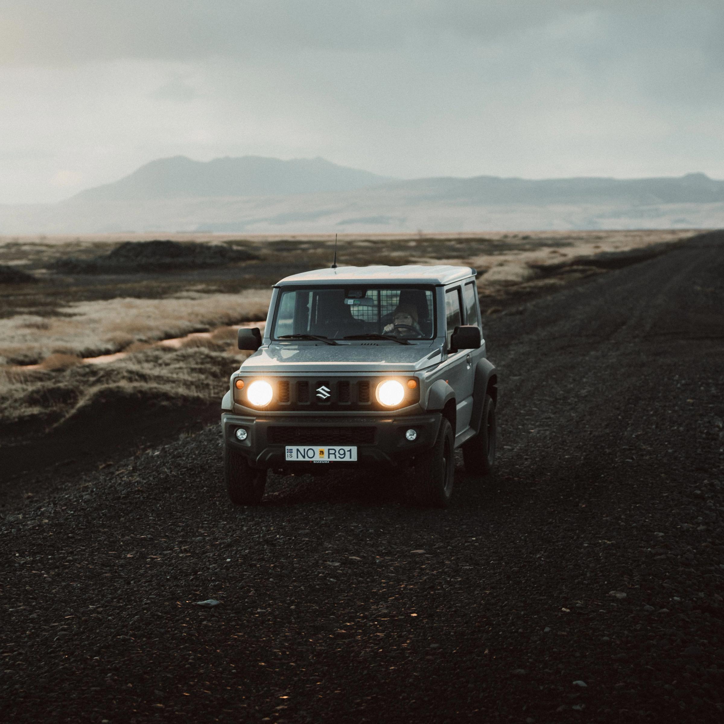 a suzuki jimny is driving down a dirt road .