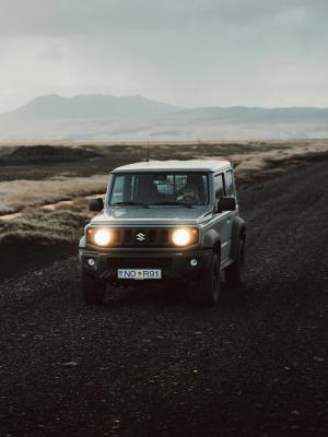A gray Suzuki Jimny with headlights on drives on a gravel road through a rugged, cloudy landscape.