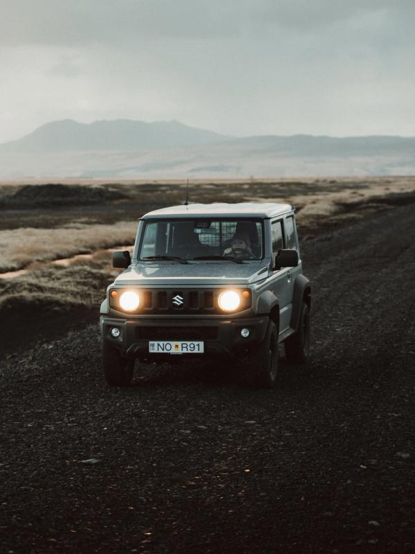 A gray Suzuki Jimny with headlights on drives on a gravel road through a rugged, cloudy landscape.