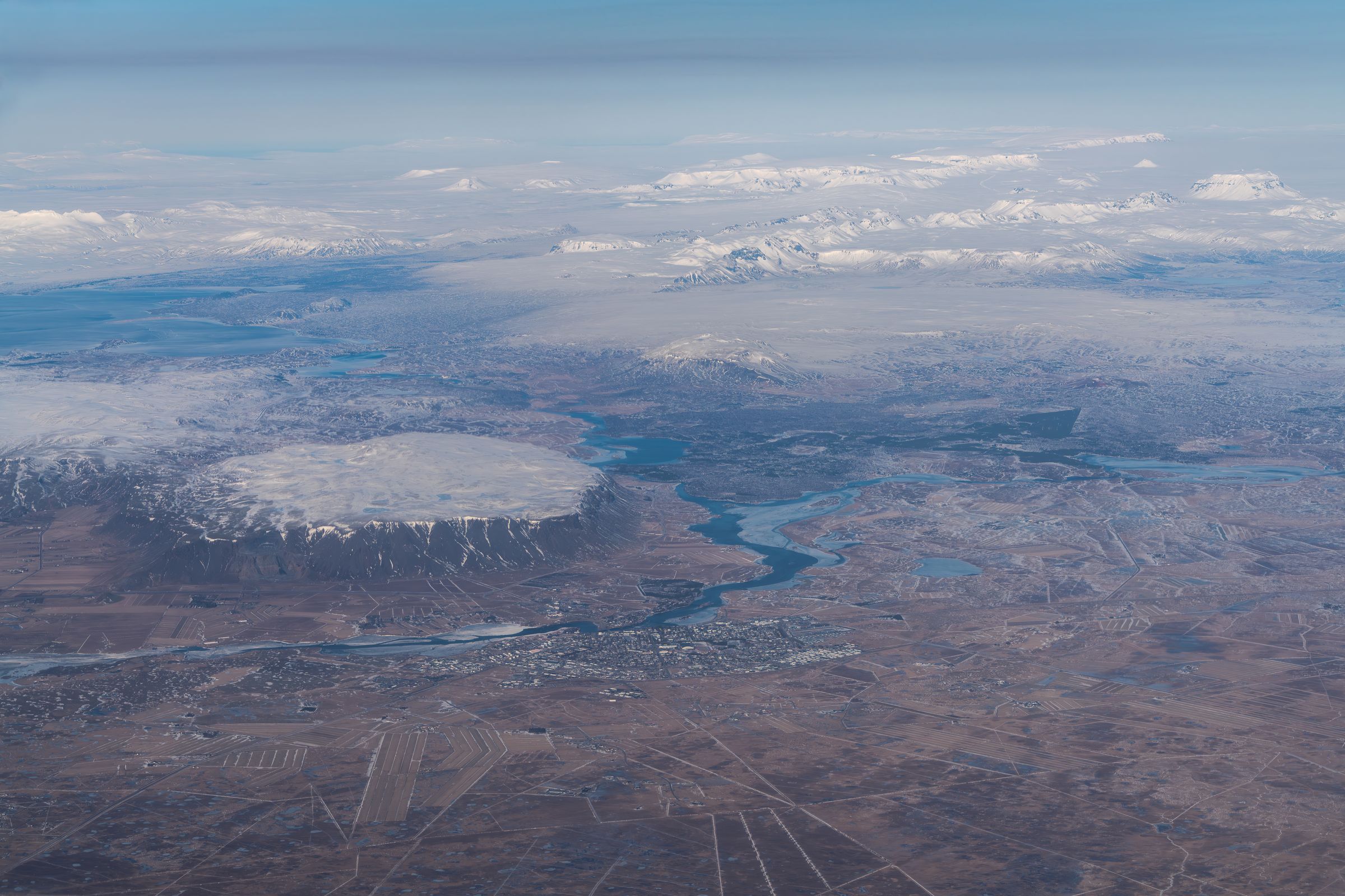 Aerial view of a vast, rugged winter landscape featuring a snow-capped, flat-topped mountain, a winding river, and distant snowy peaks under a pale sky.