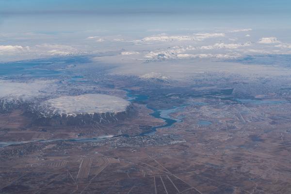 Aerial view of a vast, rugged winter landscape featuring a snow-capped, flat-topped mountain, a winding river, and distant snowy peaks under a pale sky.