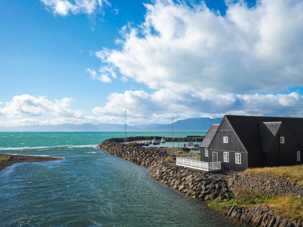 A black house on a rocky shore with a river meeting the sea, a small harbor, and mountains under a cloudy blue sky.
