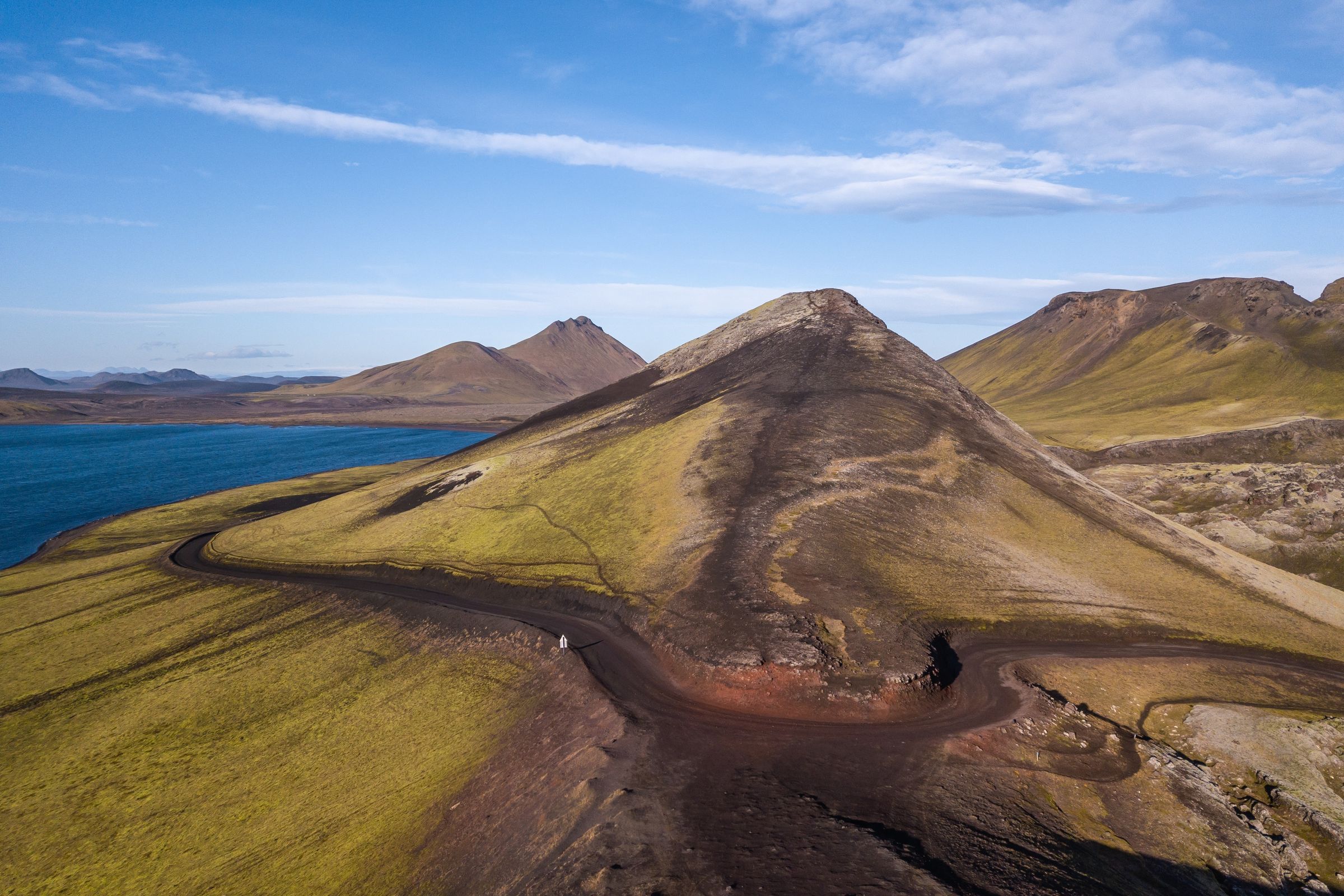Vista aérea de un paisaje volcánico escarpado con colinas verde-amarillas, una carretera oscura y sinuosa con un vehículo blanco, un lago azul y picos lejanos bajo un cielo azul.
