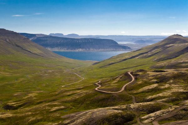 A winding dirt road curves through green valleys towards a distant fjord and mountains under a blue sky.