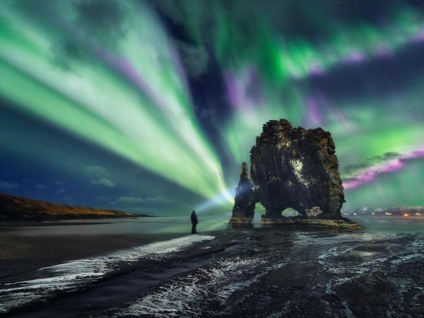 Green and purple aurora borealis over a person and a rock formation on a dark beach.