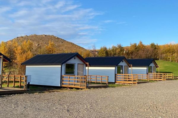 A row of white cabins with dark roofs and wooden decks, set against golden autumn trees and a rocky hill.