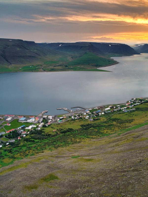 Aerial view of a coastal village on a fjord, surrounded by green hills, with a vibrant sunset sky.
