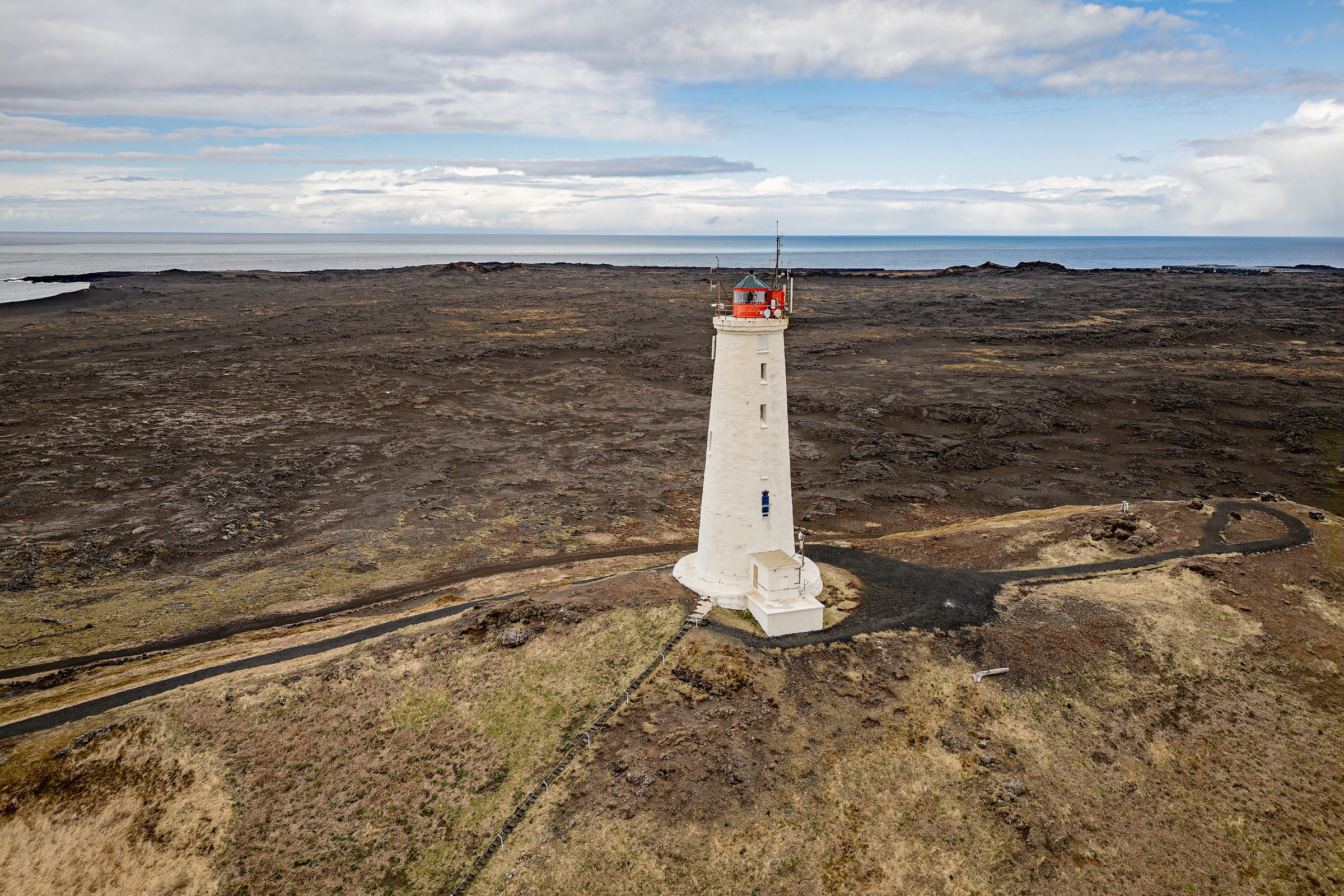 A white lighthouse with a red top on dark lava fields next to the ocean under a cloudy sky.