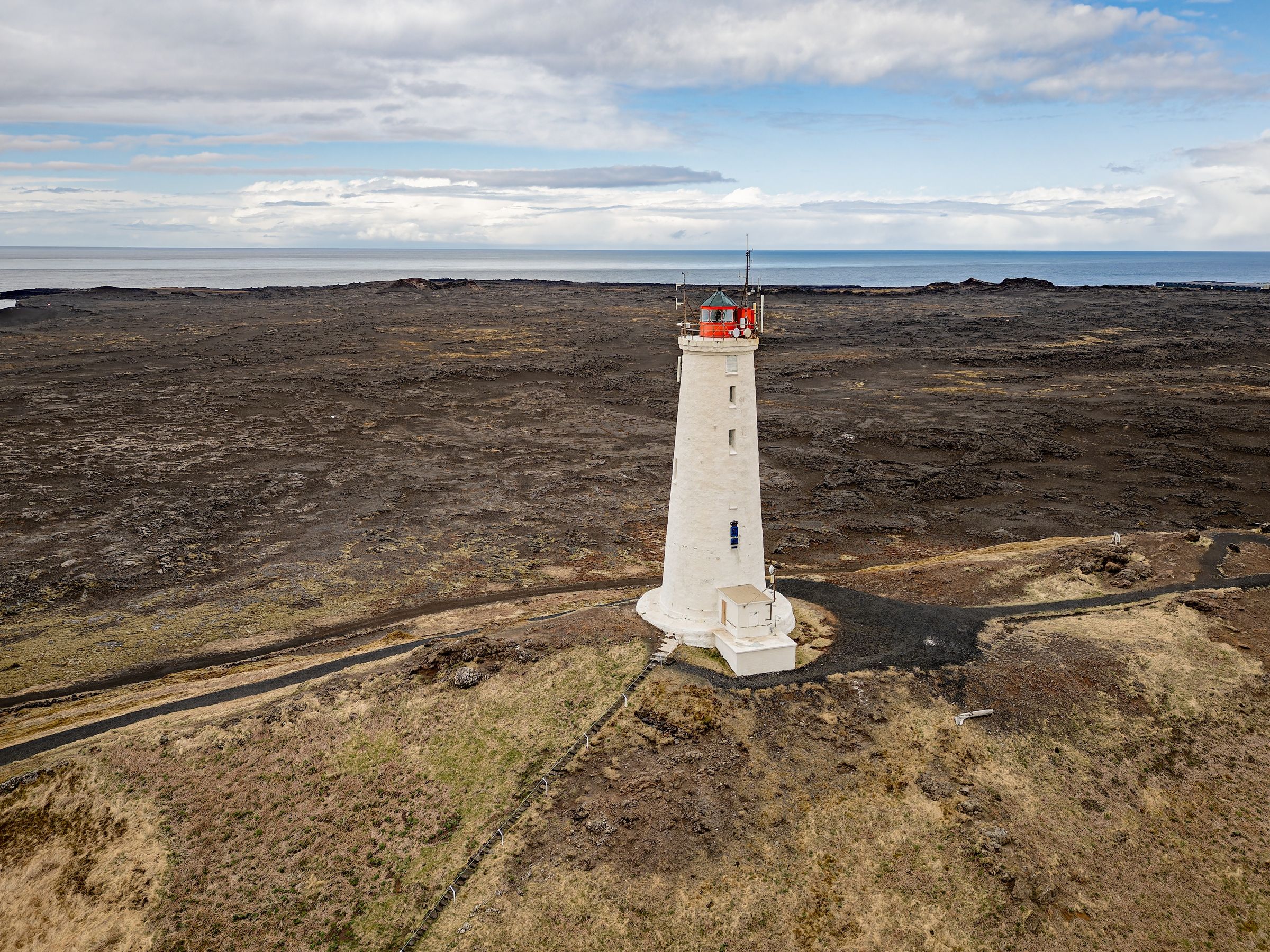 A white lighthouse with a red top on dark lava fields next to the ocean under a cloudy sky.