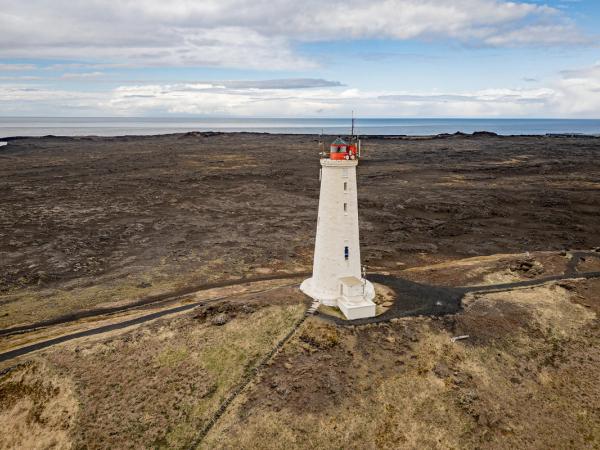 A white lighthouse with a red top on dark lava fields next to the ocean under a cloudy sky.