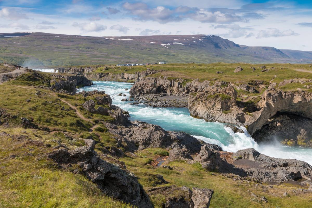 A vibrant turquoise river flows through a rocky canyon with a waterfall, green hills, and distant mountains under a blue sky.