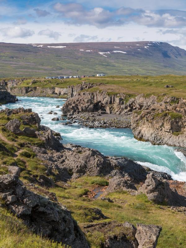 A vibrant turquoise river flows through a rocky canyon with a waterfall, green hills, and distant mountains under a blue sky.