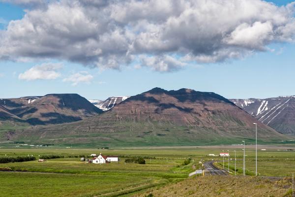Expansive green valley with a road, a small white building, and majestic, partly snow-capped mountains under a cloudy sky.