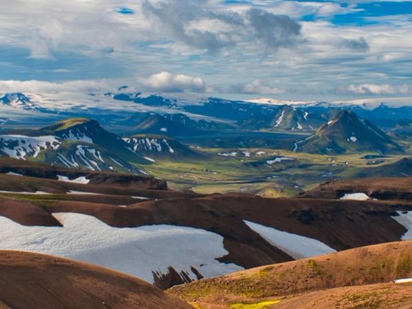 Three hikers on a snowy ridge overlooking a sweeping landscape of green valleys, distant snow-capped peaks, and a lake under a cloudy sky.