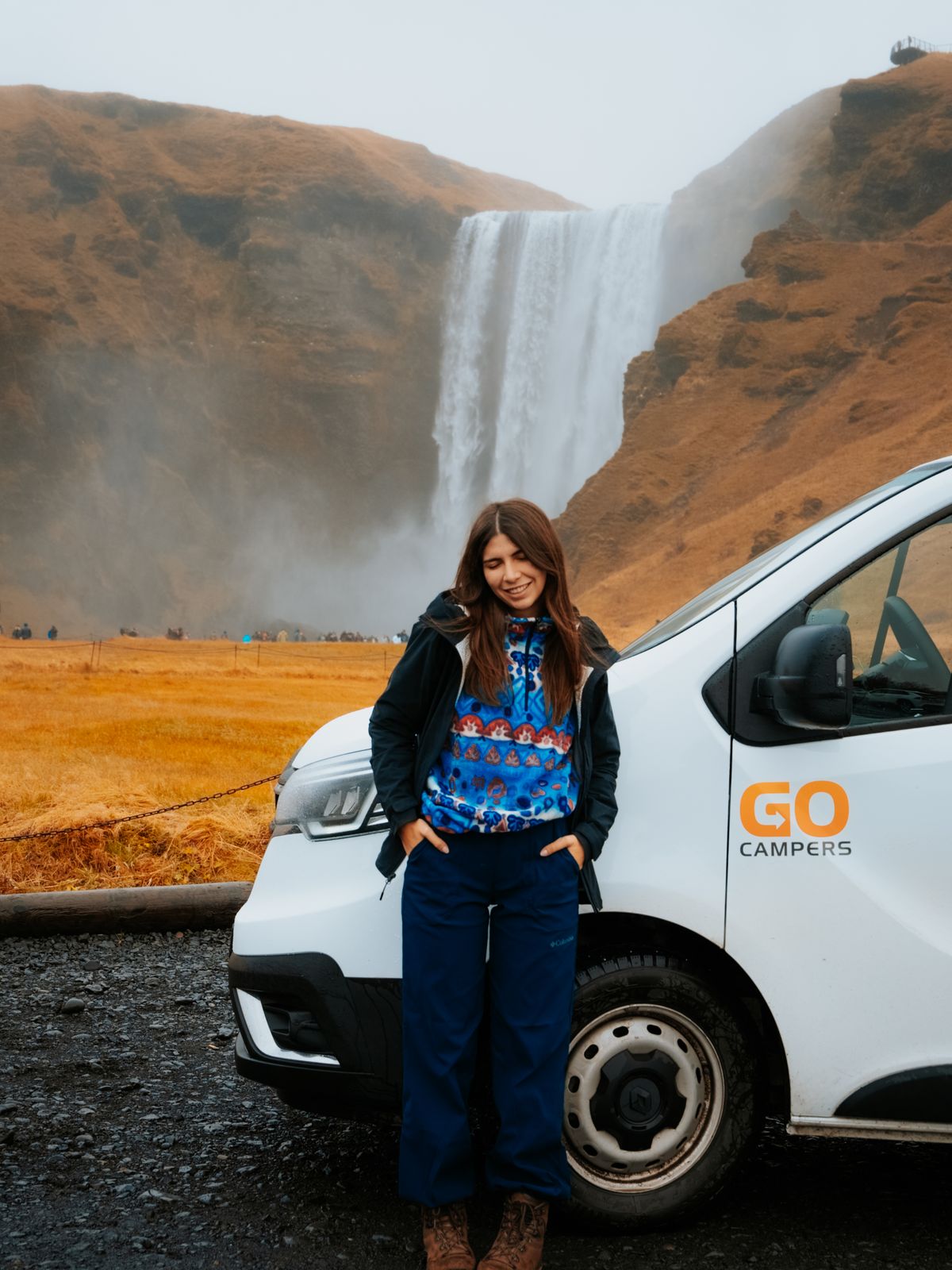Una mujer de pie junto a una furgoneta blanca frente a una cascada.