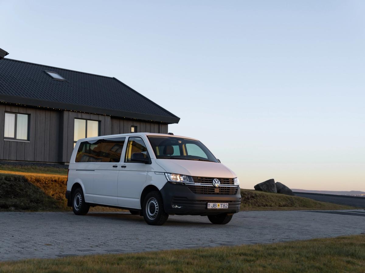 Volkswagen Caravelle – Vorderansicht vor einem Haus Volkswagen Caravelle Mietwagen in Island, geparkt in einer Einfahrt neben einem modernen Haus mit Grasdach