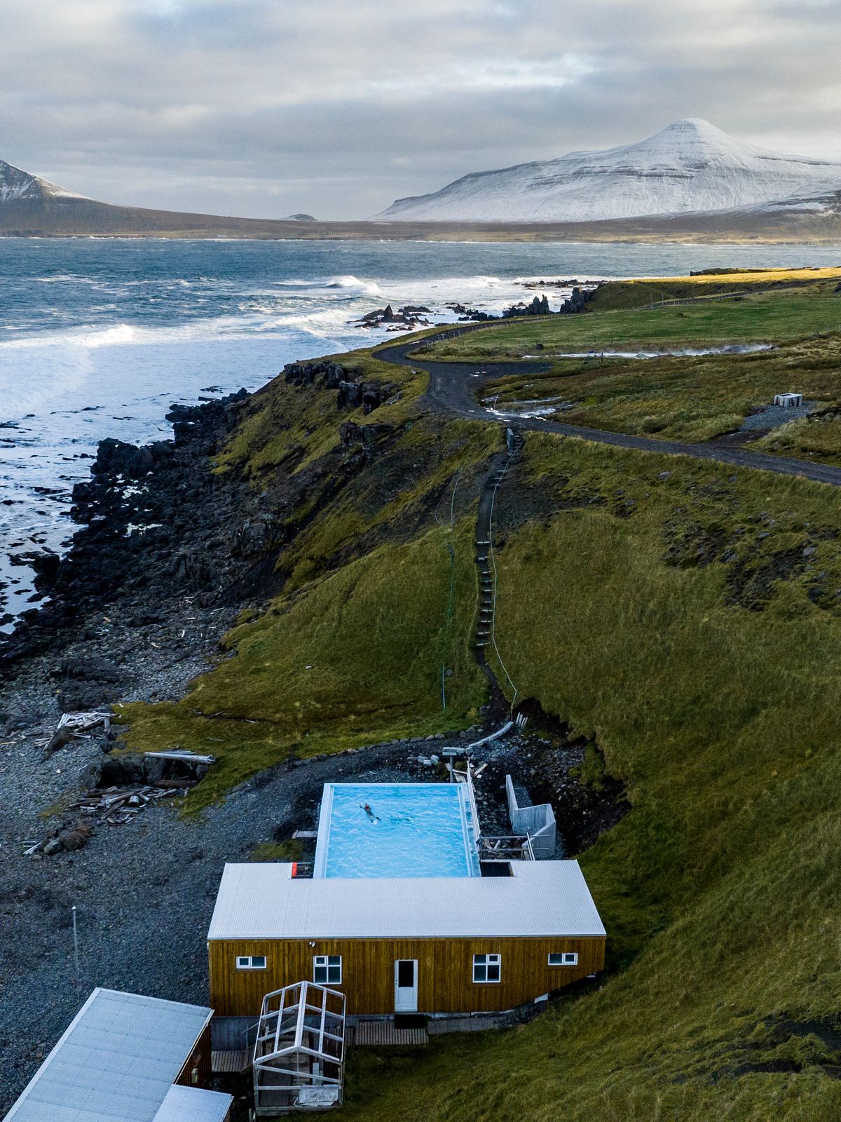 A building with a bright blue outdoor pool on a rugged, rocky coast with breaking waves, a winding dirt road, green hills, and distant snow-capped mountains under a cloudy sky.