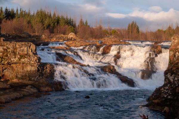 A multi-tiered waterfall cascades over brown rocks into a river, with evergreen trees in the background.