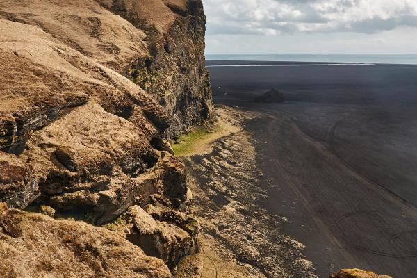 A rugged cliff covered in dry, golden-brown grass overlooks a vast black sand beach and the ocean under a cloudy sky.