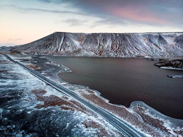 Aerial view of a road alongside a dark lake, with a snow-dusted, textured mountain under a pale twilight sky.
