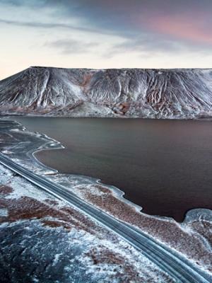 Aerial view of a road alongside a dark lake, with a snow-dusted, textured mountain under a pale twilight sky.