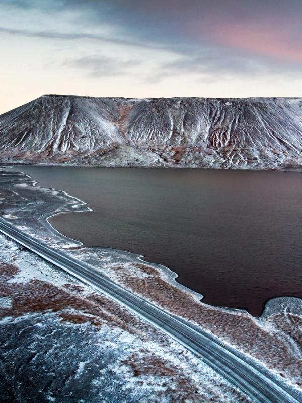 Aerial view of a road alongside a dark lake, with a snow-dusted, textured mountain under a pale twilight sky.