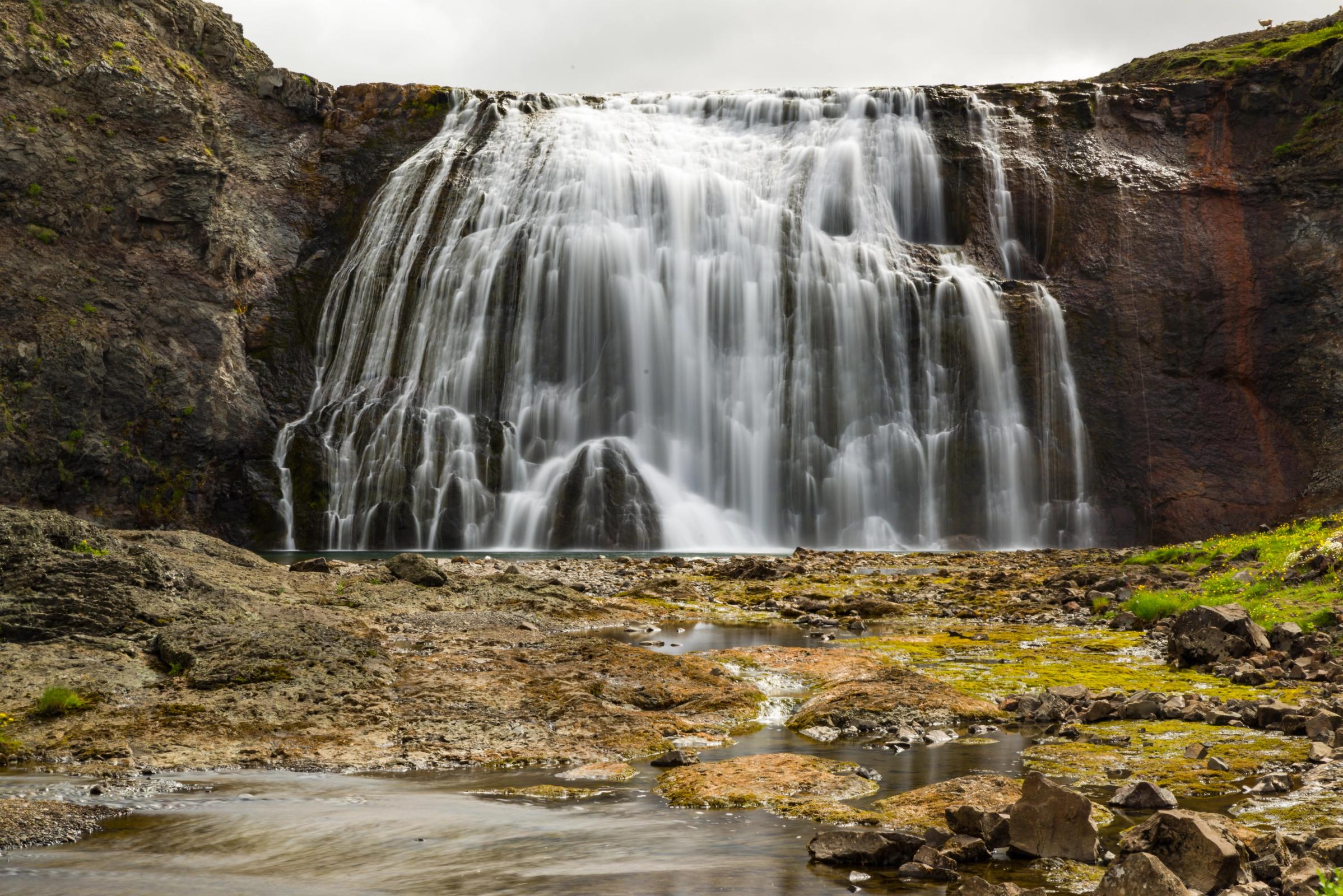 A wide waterfall with silky white water cascades over dark rocky cliffs into a stony, mossy streambed.