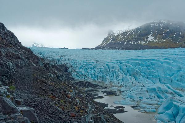A vibrant blue glacier with deep crevasses alongside a dark, rocky mountainside under a cloudy sky.