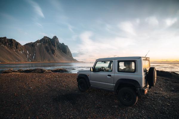 A silver SUV parked on a rocky beach with dramatic mountains and the ocean at sunset.