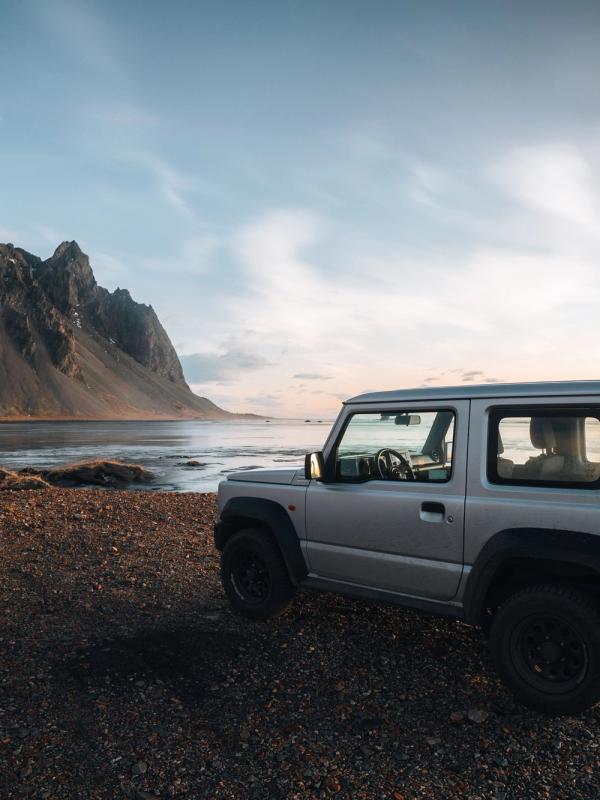 A silver SUV parked on a rocky beach with dramatic mountains and the ocean at sunset.