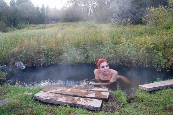 A woman with red hair relaxes in a steaming outdoor hot spring surrounded by tall grass.