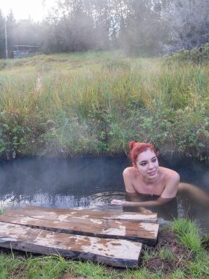 A woman with red hair relaxes in a steaming outdoor hot spring surrounded by tall grass.