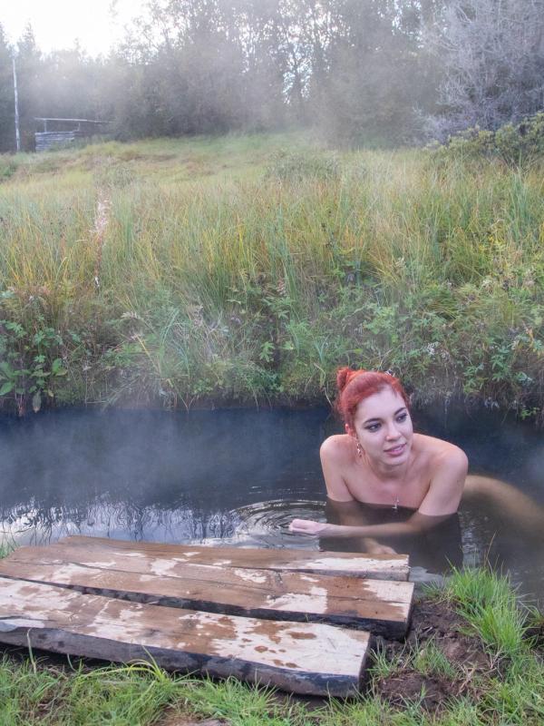 A woman with red hair relaxes in a steaming outdoor hot spring surrounded by tall grass.
