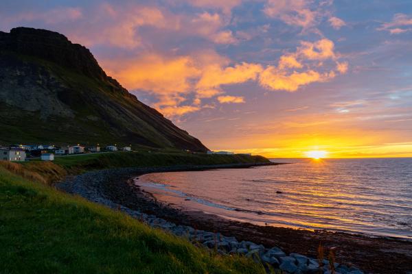 A vibrant orange and pink sunset over a coastal village at the base of a green mountain, with a rocky beach and ocean.