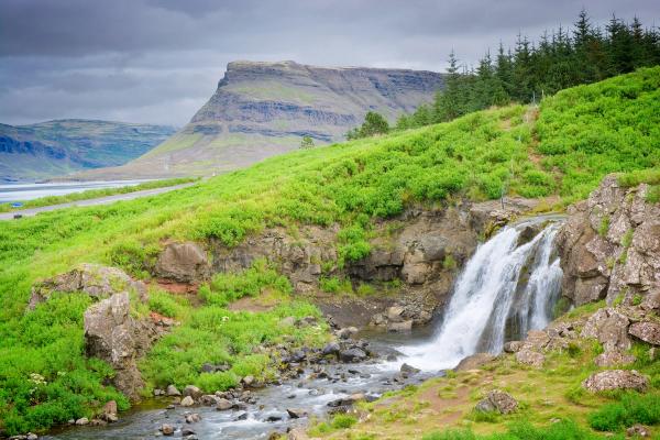 A waterfall cascades down rocky terrain amidst vibrant green hills, with a large mountain and cloudy sky in the background.