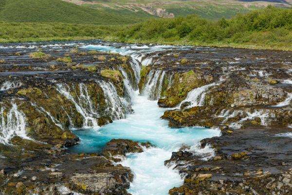 Vibrant blue waterfalls cascade over dark, mossy rocks through a green landscape.