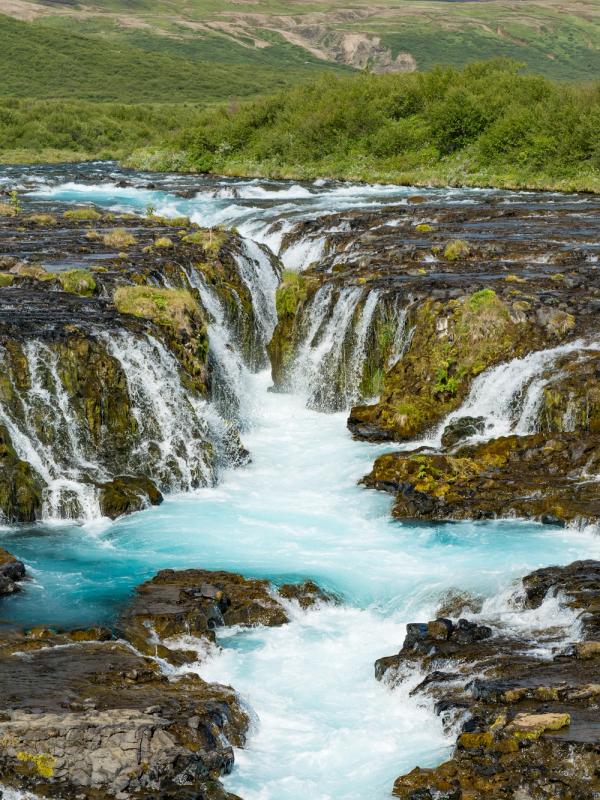 A bright blue glacial river cascades over dark, rugged rocks, framed by green hills.